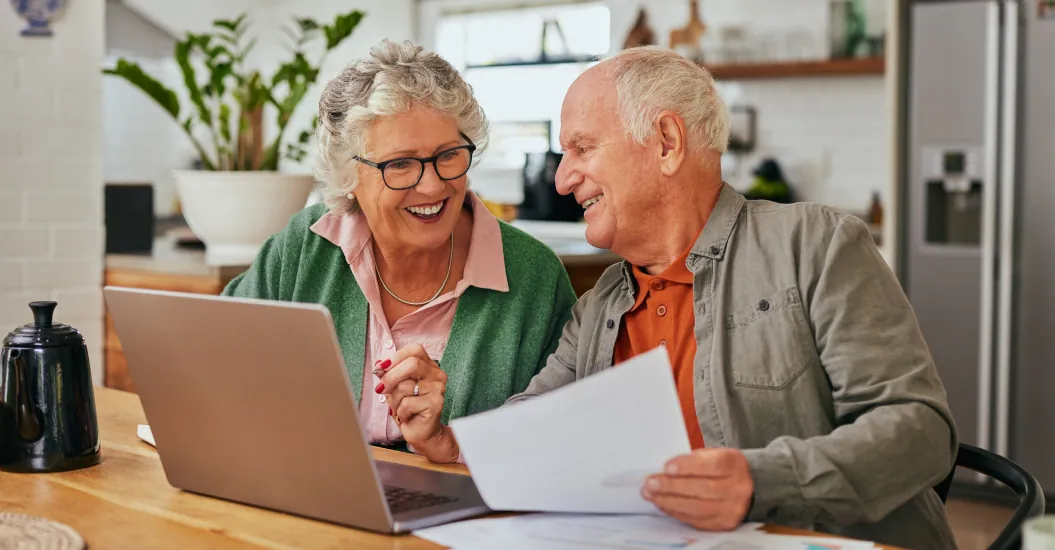 Two older adults sit at kitchen table, smiling and looking at the savings they are making on their insurance in front of an open laptop.