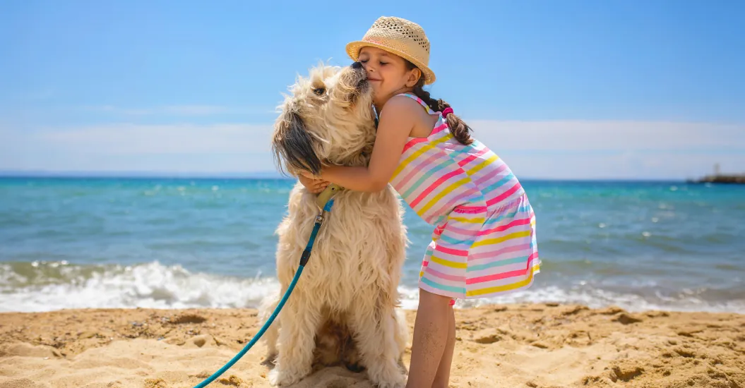 A young girl on holiday in a striped dress and straw hat hugs a large, fluffy dog on a sandy beach, with the ocean and blue sky in the background.