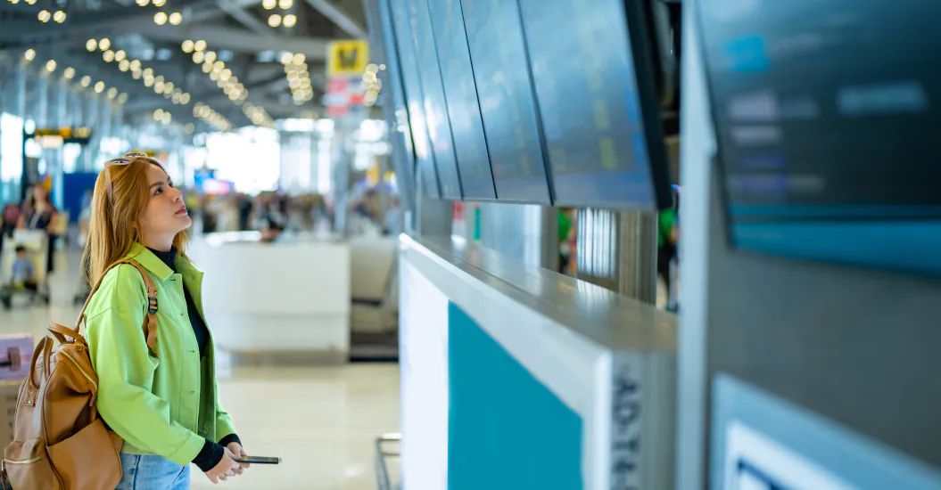 A woman with a backpack looks up at flight information screens in a busy airport terminal.