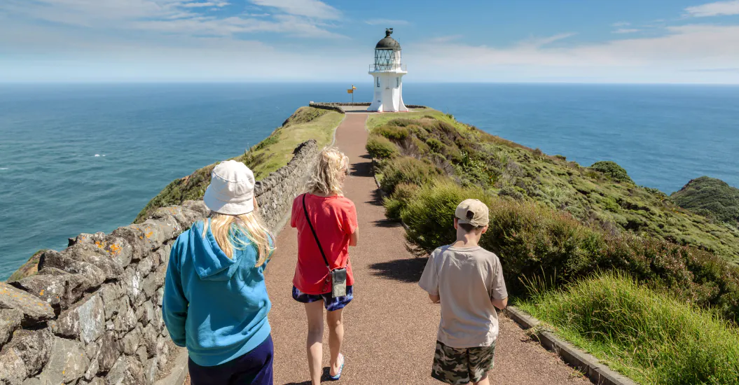 Three people walk along a path towards the Cape Reinga lighthouse situated on a grassy cliff overlooking the ocean