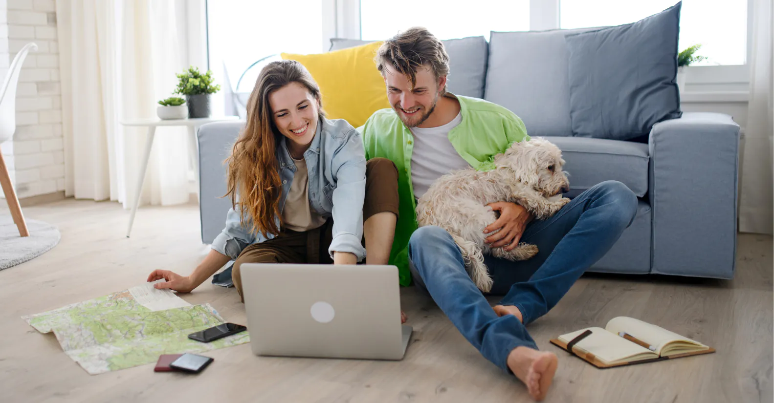 A couple sits on the floor by a laptop, with a dog on one person’s lap, maps and notebooks nearby, planning or researching in a bright living room.